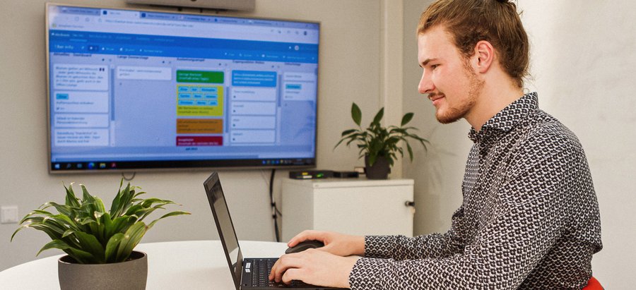 Bachelor of Science - Verwaltungsinformatik E-Government. Foto: Thomas Grimm Ein Mann sitzt an einem modernen Schreibtisch und bearbeitet etwas am Laptop. Im Hintergrund ist ein Kanban-Board auf einem Bildschirm zu sehen. Foto: Thomas Grimm