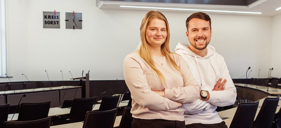 Zwei junge Personen stehen angelehnt im Sitzungssaal neben dem Kreis Soest-Logo. Foto: Thomas Grimm