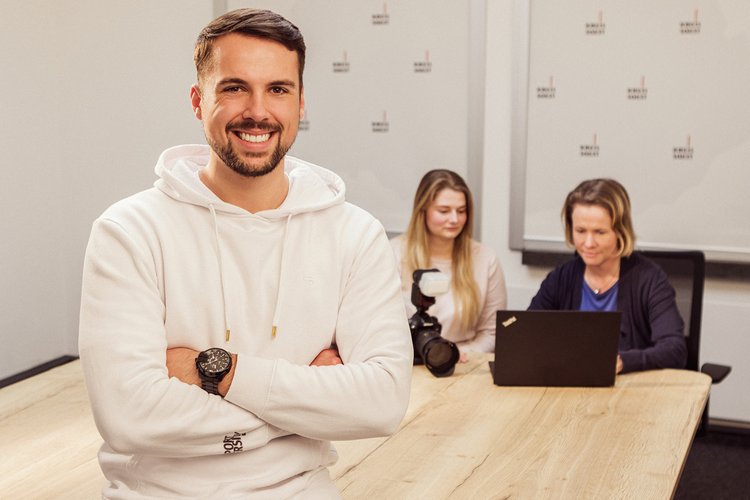 Volontär. Foto: Thomas Grimm Ein Mann steht vor einem Besprechungstisch, an dem zwei Frauen auf einen Laptop schauen. Foto: Thomas Grimm