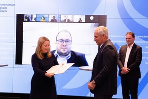 Ein Förderbescheid wird an eine Person übergeben. Im Hintergrund ist auf einer Leinwand ein Foto von dem Mobilfunkkoordinator des Kreises Soest zu sehen. Foto: Bundesministerium für Digitales und Verkehr (BMDV)
